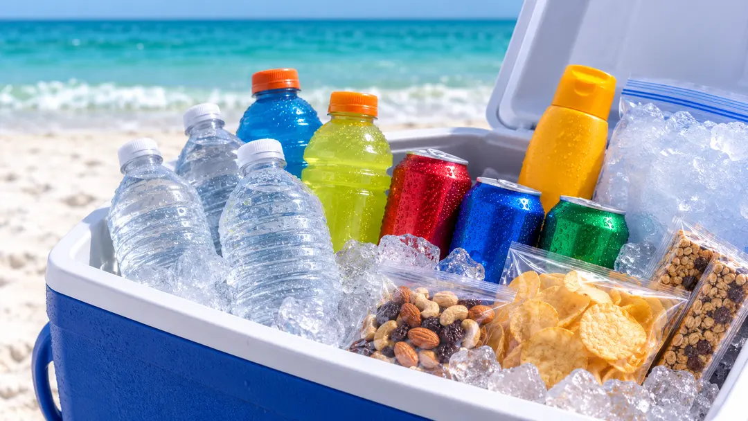 A cooler of iced water, sports drinks, snacks and sunscreen on the sand.